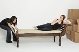 Young Asian woman pulls her room mate accross the room on a lounging sofa.  The lazy room mate pretends to be tired.