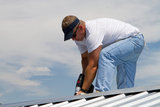 Construction worker uses a power drill to attach a cap the the top of a sheet metal roofing job with screws.